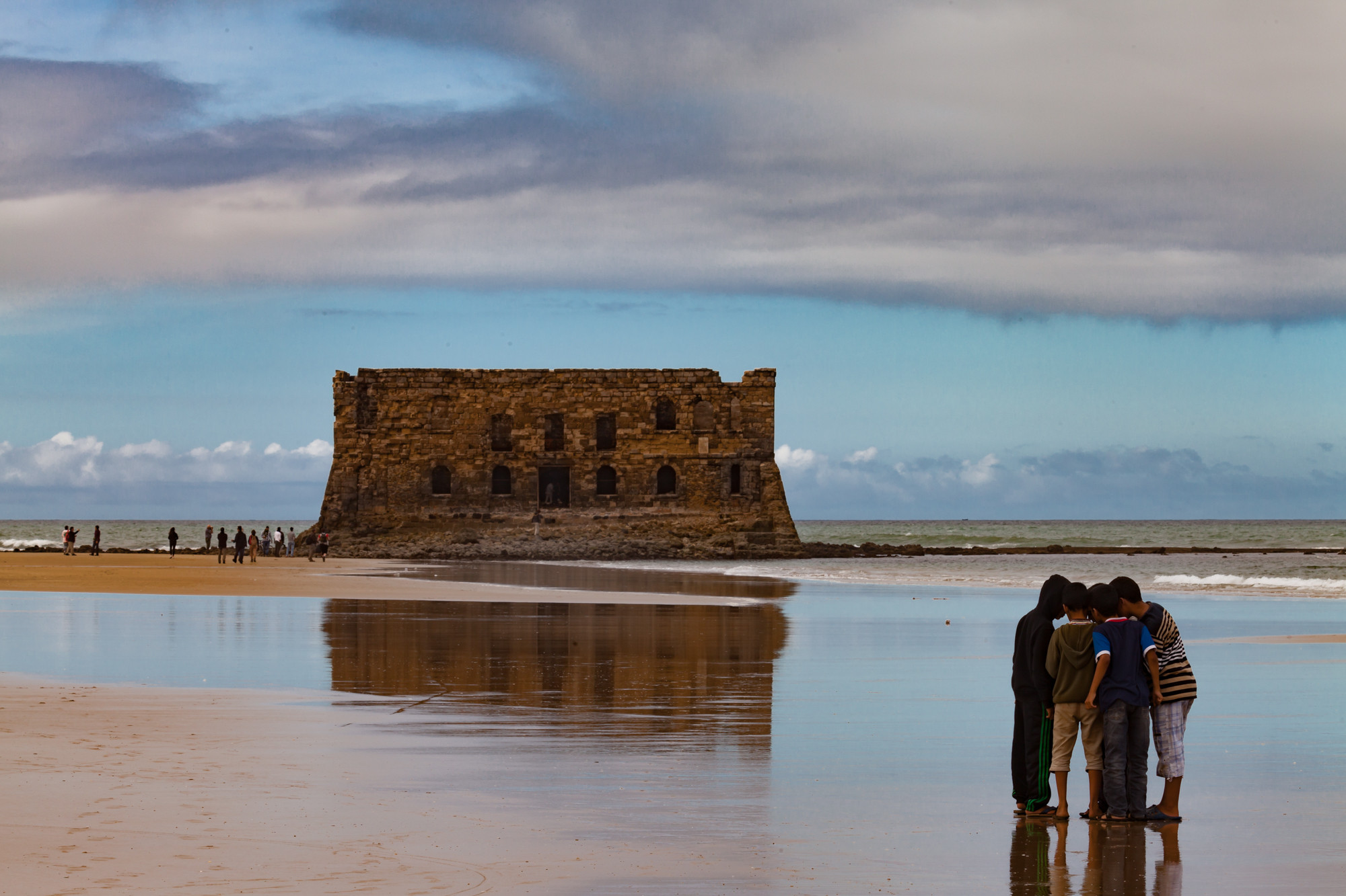 Grupo de personas reunido en una playa frente a un edificio de piedra