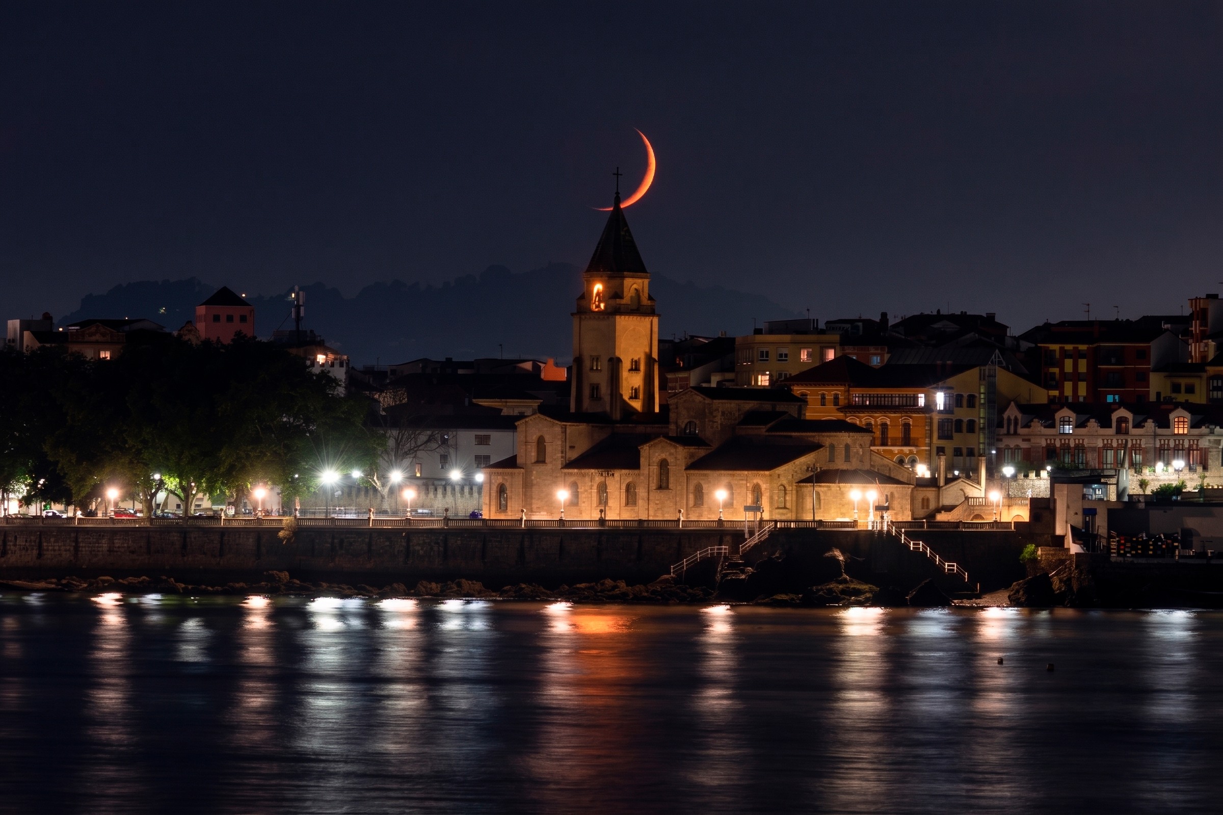 Luna creciente sobre una localidad costera iluminada al anochecer, fotografía de Chechu Senís